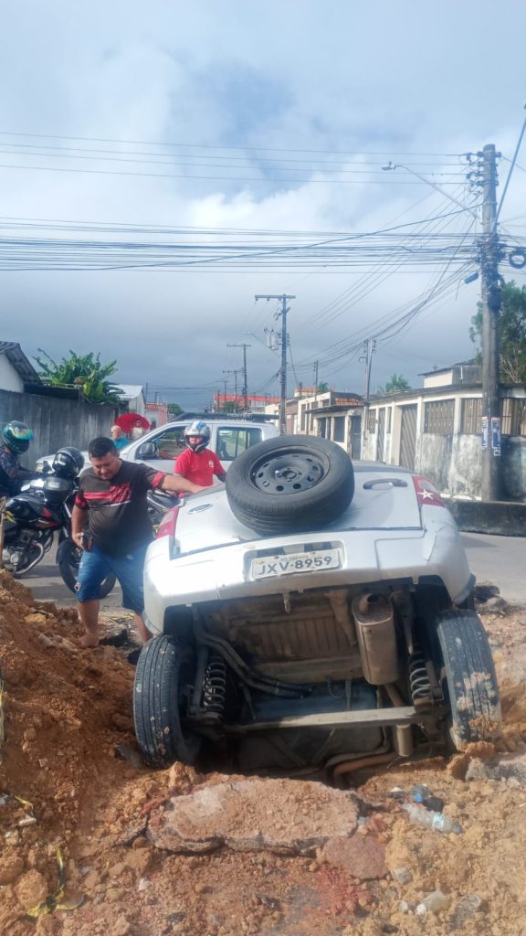 Carro de passeio parcialmente submerso dentro de uma cratera aberta em rua de Manaus durante obras de drenagem profunda.