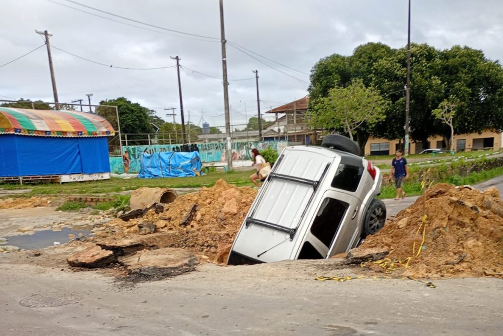 Carro de passeio parcialmente submerso dentro de uma cratera aberta em rua de Manaus durante obras de drenagem profunda.