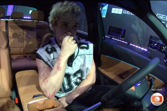 Juliano comemorando a primeira vaga na final do Big Brother Brasil 26 ao lado de um carro zero.