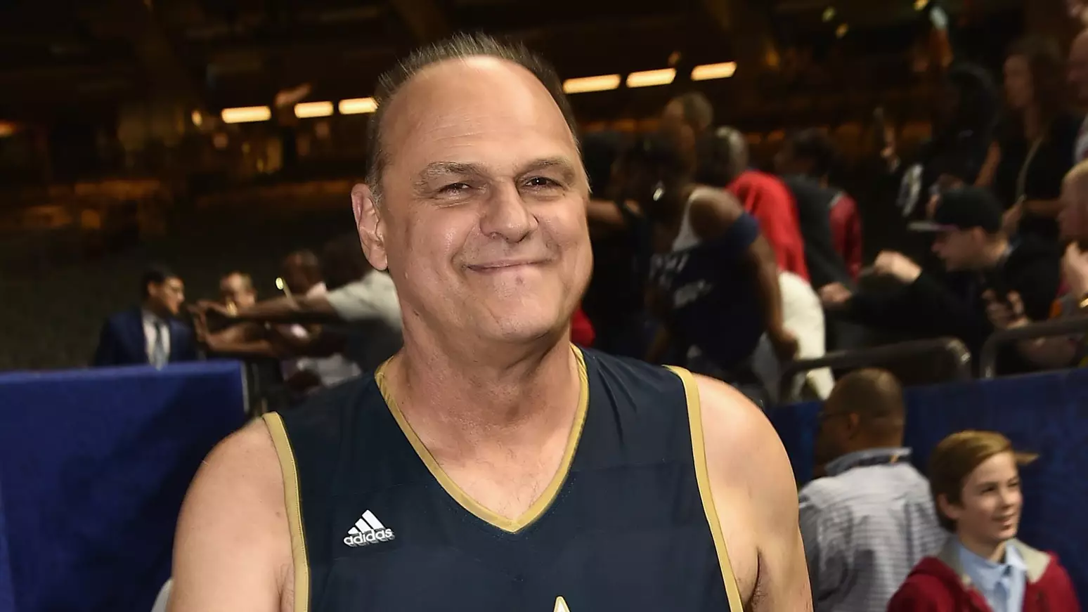 Oscar Schmidt sorridente em evento oficial de basquete com iluminação cênica ao fundo.