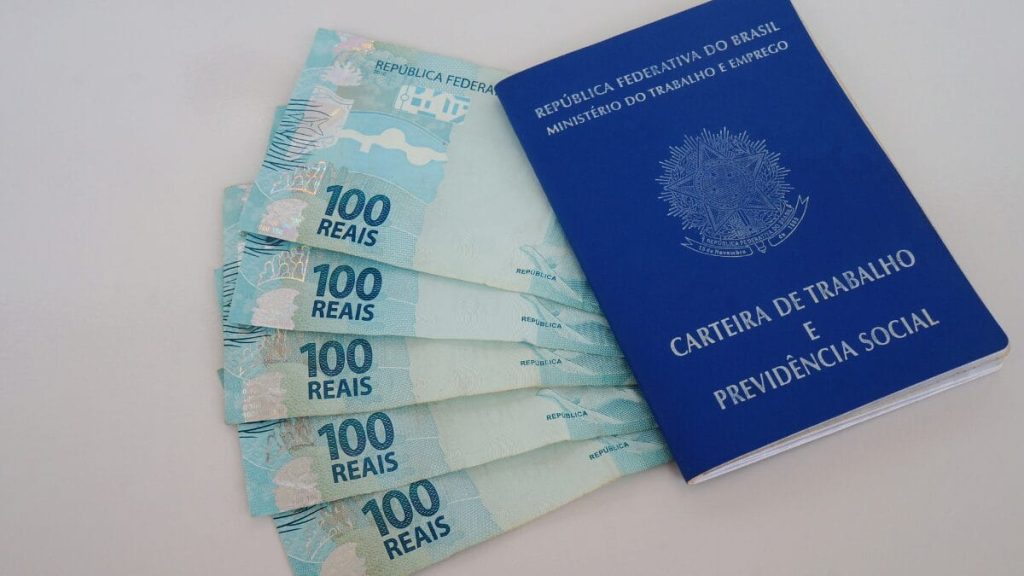 Trabalhador segurando carteira de trabalho e cédulas de dinheiro sobre o abono salarial pis pasep